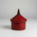 A small round wooden box with a matching pointed lid, painted in a glossy dark red finish, set on a dark tabletop against a white wall.