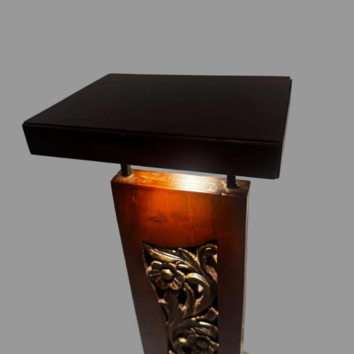 A dark brown wooden display stand or pedestal with a rectangular top and an ornate carved floral panel on the front, photographed indoors against a plain wall.